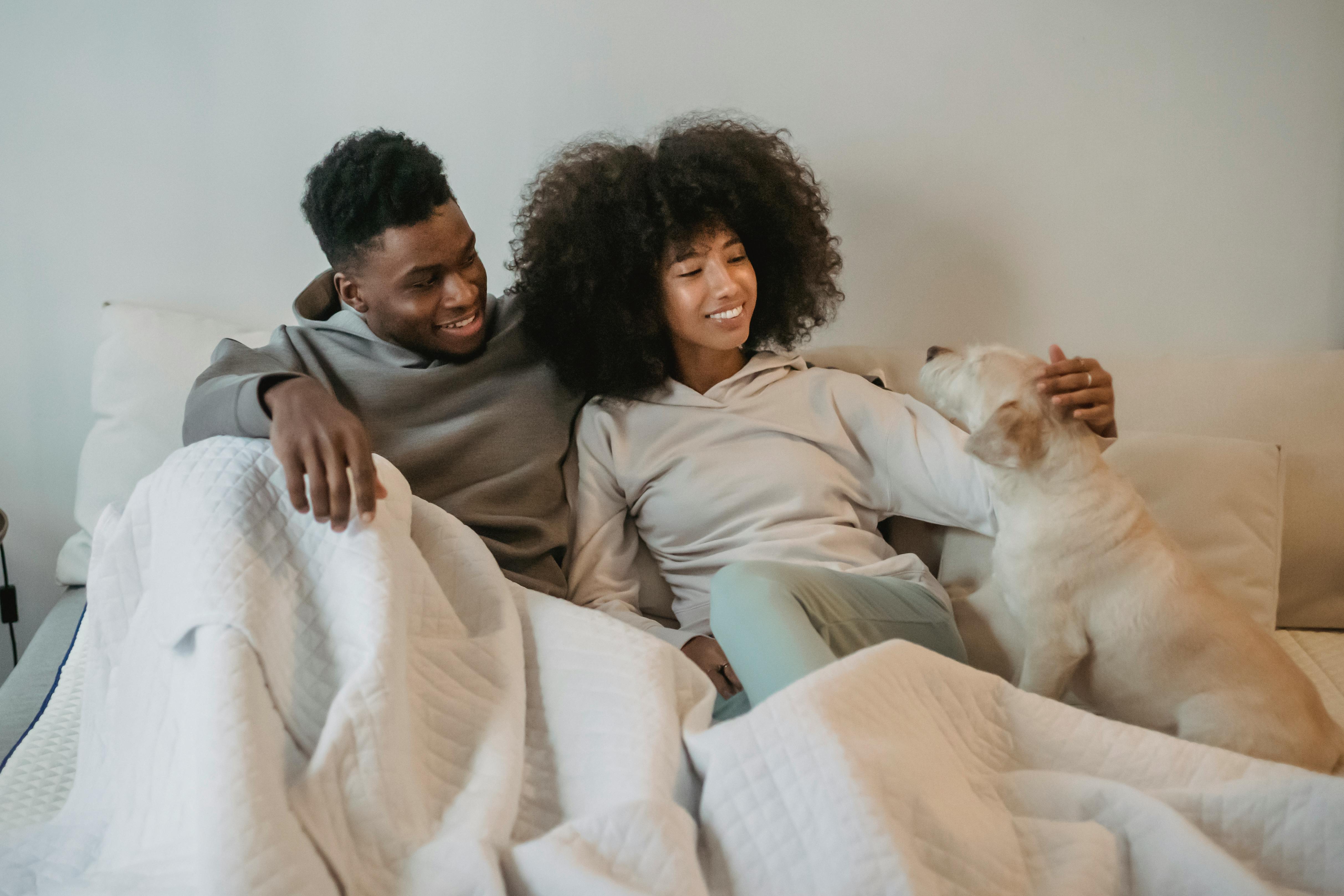 A Black couple relaxes in bed, looking fondly at their dog and patting its head.